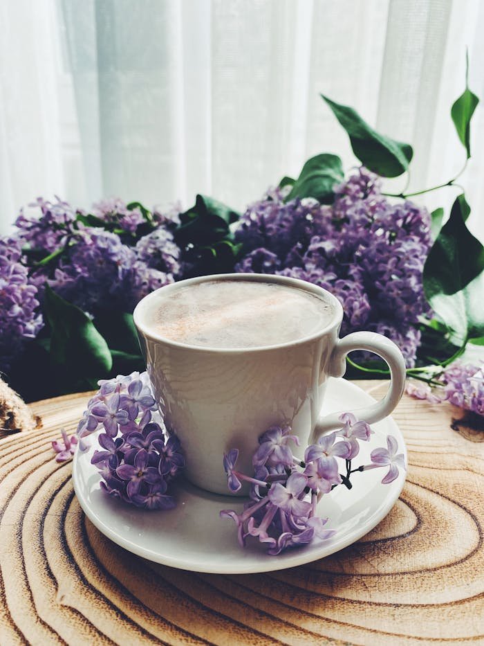 about-03 A serene cup of coffee surrounded by lilac blossoms on a rustic wooden table, perfect for a calming morning setting.