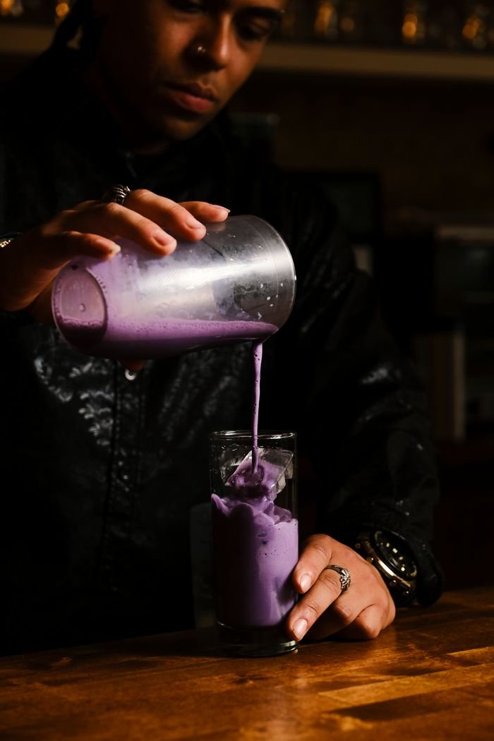 Mastering the First Impression: Your intriguing post title goes here Close-up of a bartender pouring a vibrant purple cocktail in a dimly lit bar.