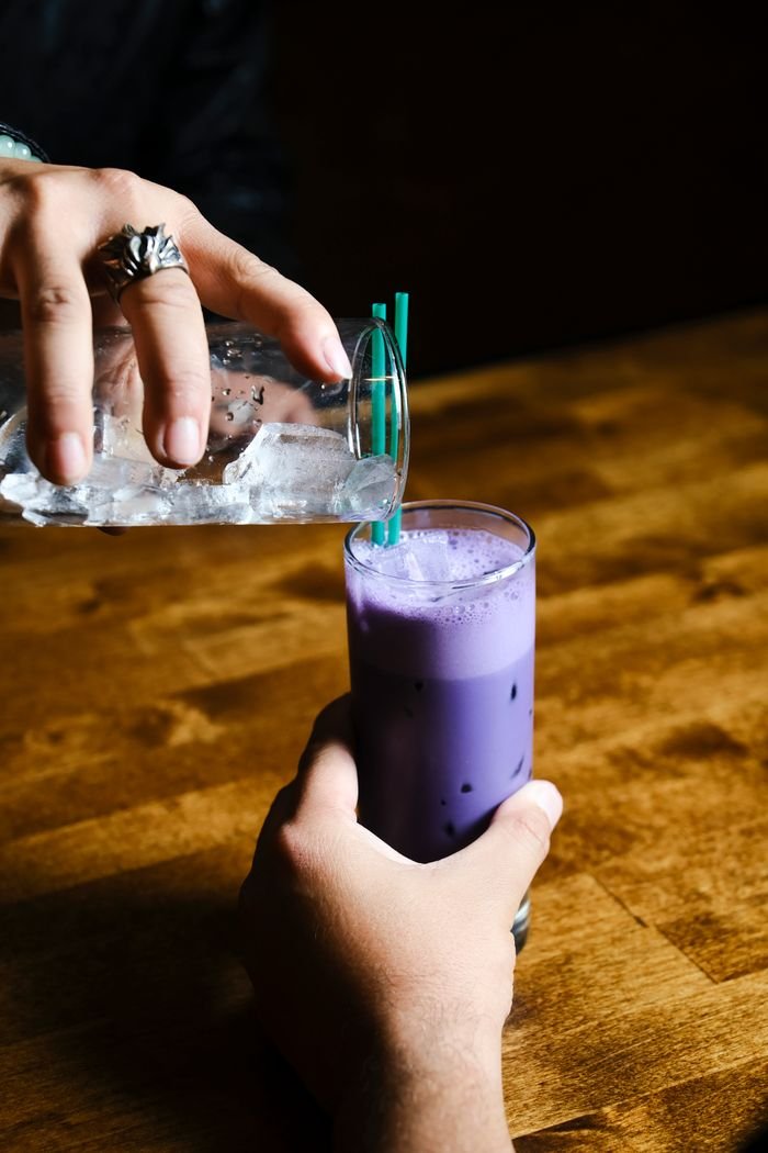 project-02-a Close-up of hands pouring ice water into a glass with purple drink on a wooden table.