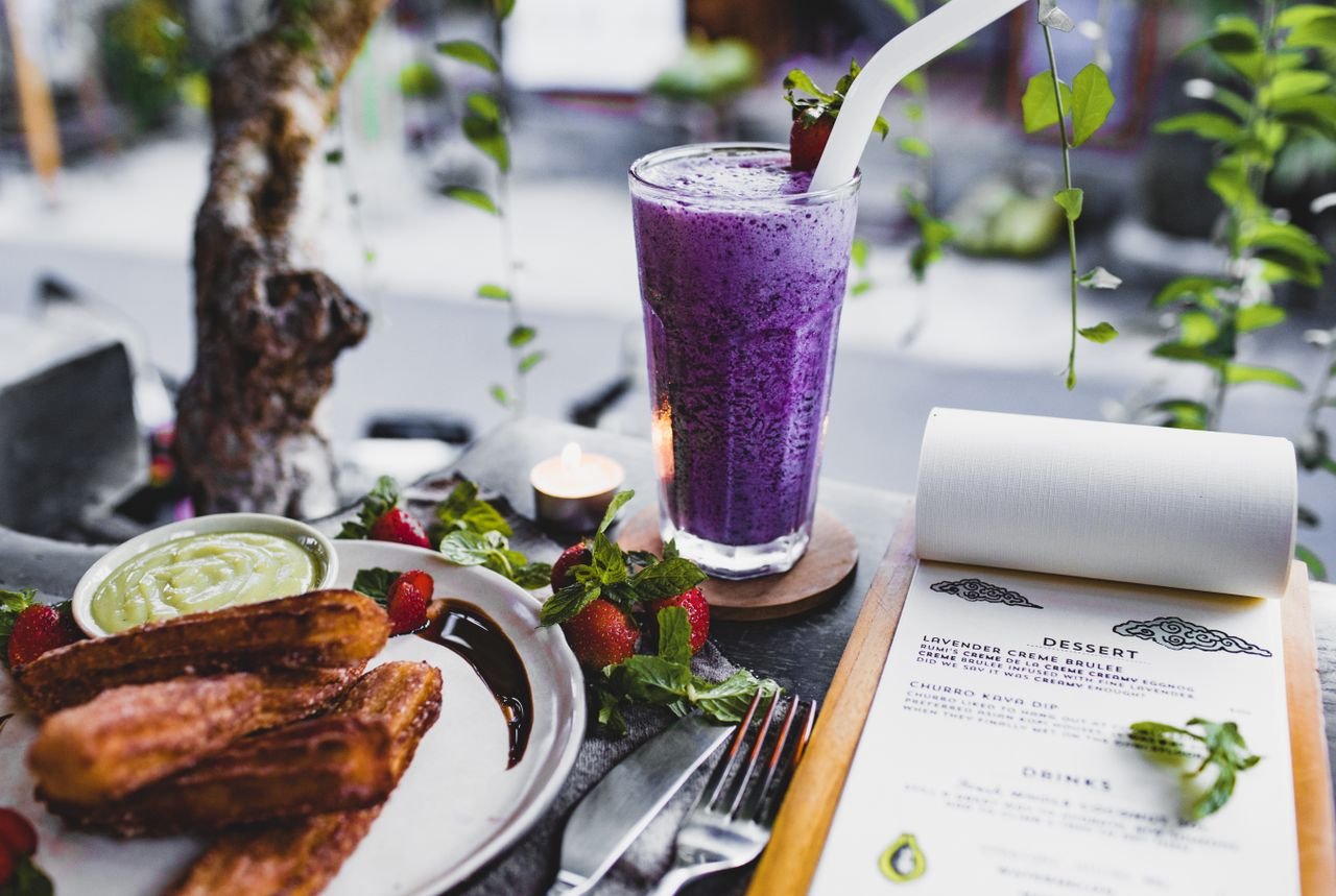 From above of glass of lavender creme brulee garnished with strawberry and plate of churro with avocado sauce placed on table with knife and fork and menu and candle against window decorated with plants