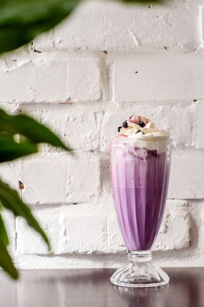 about-header Purple blueberry milkshake with whipped cream against a rustic white brick wall.